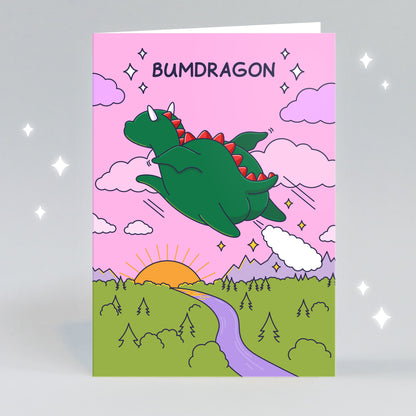 A funny greeting card of a dragon farting as he files over a beautiful sunset with a pink sky. The caption reads 'Bumdragon'.