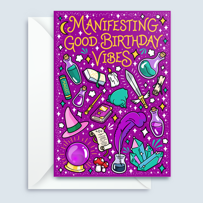 A purple birthday card with whimsical witch-themed illustrations and the text 'Manifesting Good Birthday Vibes,' shown with a white envelope on a light gray background.