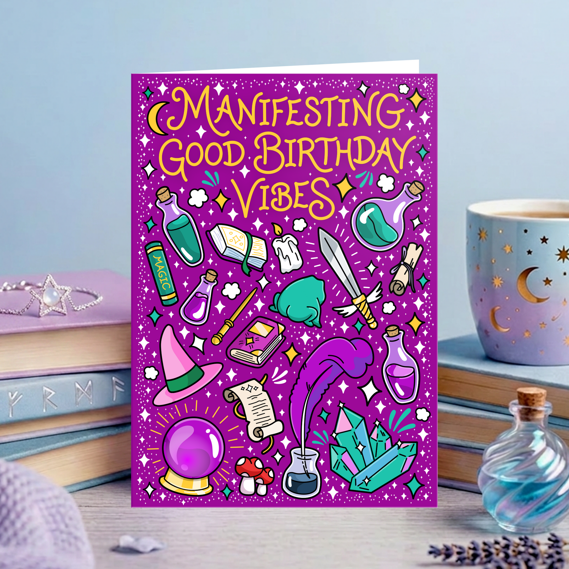 A rectangular greeting card with a purple glittery background and colorful illustrated magic-themed graphics, standing upright on a tabletop. The card reads “MANIFESTING GOOD BIRTHDAY VIBES” in yellow lettering.
