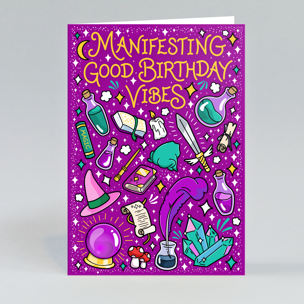 A rectangular greeting card standing upright on a light gray background. The card has a bright purple glitter-style background with the phrase “MANIFESTING GOOD BIRTHDAY VIBES” in large yellow lettering at the top, surrounded by whimsical illustrated potion bottles, crystals, a wand, a hat, a potion bottle, mushrooms, and sparkles.