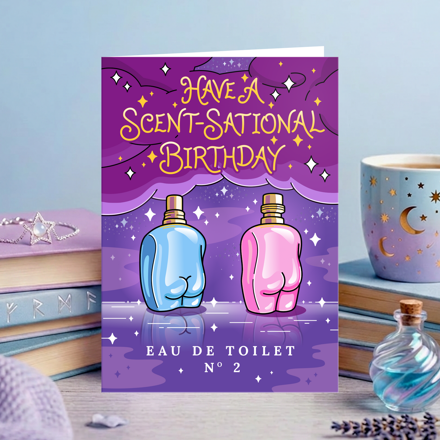 A greeting card featuring a purple and blue illustrated background with two rear-view perfume bottles and the text 'HAVE A SCENT-SATIONAL BIRTHDAY' and 'EAU DE TOILET N° 2'. The card is displayed upright on a tabletop surrounded by books, a cup, and decorative items.