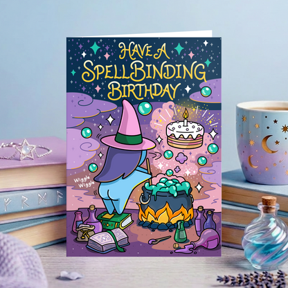 A greeting card displayed on a tabletop with books, a cup, and small decorative items. The card reads “HAVE A SPELLBINDING BIRTHDAY” in large yellow letters over a whimsical illustration of a witch in a pink hat standing by a cauldron with a cake floating above.