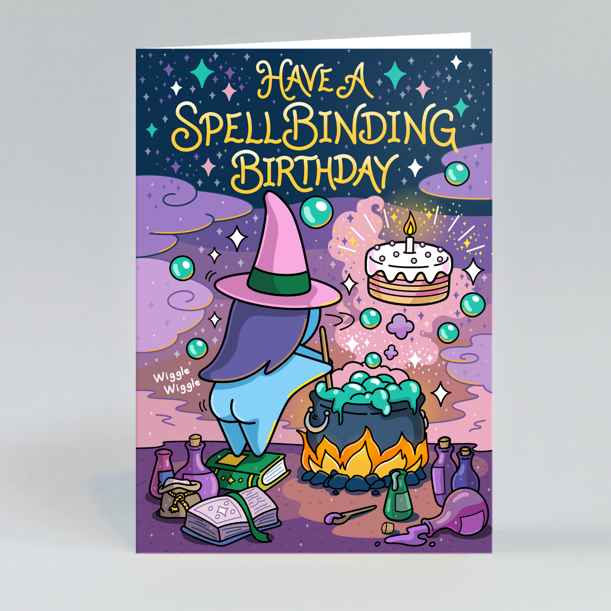 A greeting card featuring a whimsical illustration of a witch in a pink hat casting a spell with a cauldron, potion bottles, stars, and a birthday cake. The card reads 'HAVE A SPELLBINDING BIRTHDAY' in large yellow letters against a dark blue starry sky.