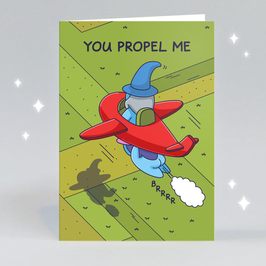 A greeting card of a Wizard sitting in a cute red plane with a Witch flying propelling him forward. The caption reads 'You Propel Me'.